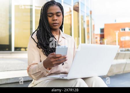 Una donna d'affari concentrata utilizza il suo smartphone mentre lavora al suo laptop, seduto all'aperto in una giornata di sole, forse in una città come Barcellona durante la S. Foto Stock