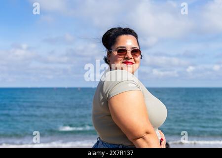 Una donna gioiosa che indossa gli occhiali da sole si trova in riva al mare in una giornata estiva di sole a Barcellona, trasudando appagamento e fiducia Foto Stock