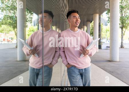 Uno studente asiatico con una felpa rosa tiene i documenti all'aperto, sorridendo mentre guarda lontano dalla fotocamera, con un riflesso chiaro in un vetro pa Foto Stock