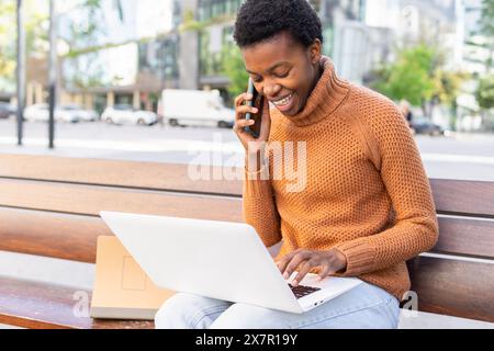 Una donna afro-americana sollevata esegue il multitasking sul suo computer portatile mentre parla su uno smartphone, seduto su una panchina all'aperto con un'espressione gioiosa. Foto Stock