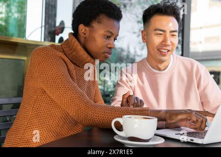 Una giovane donna e un uomo si siedono a un tavolo in un bar, la donna in un dolcevita guarda con attenzione lo schermo di un notebook mentre l'uomo in una felpa rosa si sta smontando Foto Stock