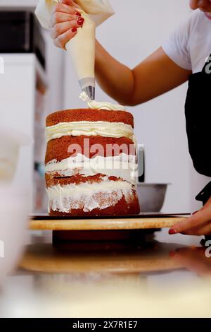 Una persona applica la glassa di panna su una torta di spugne multistrato con un sacchetto di tubature in un ambiente di cucina Foto Stock
