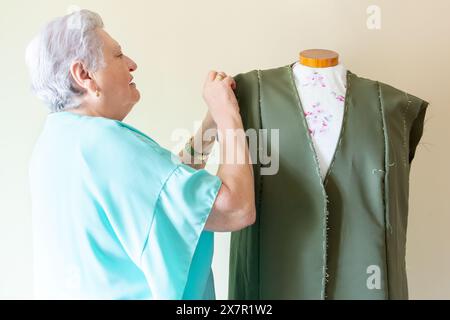 Una donna anziana indossa meticolosamente un abito verde su un manichino, focalizzato sul compito da svolgere in un laboratorio di cucito Foto Stock