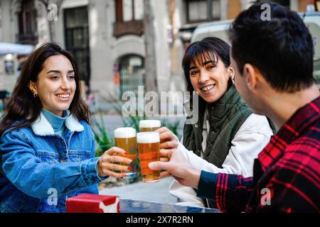 Gli amici LGBTQ si godono una riunione informale all'aperto, brindando con delle birre e condividendo le Risate, con due donne che si guardano l'un l'altra e un uomo che guarda lontano. Foto Stock