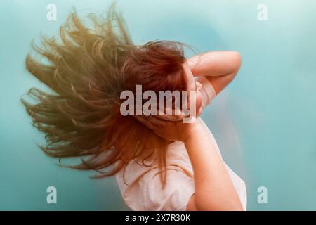 Una giovane donna in angoscia tiene la testa, i capelli che scorrono, adagiati su un sereno sfondo di tè, illustrando i sintomi dell'ipoglicemia Foto Stock