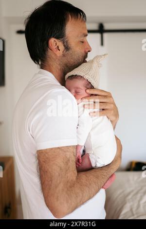 Un padre tiene con amore il suo neonato, che indossa un berretto a maglia, in un caldo abbraccio a casa Foto Stock