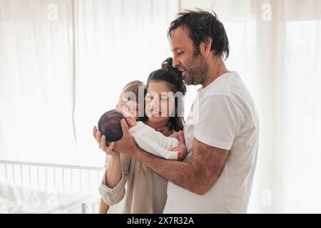Una famiglia felice con un padre, una madre e una figlia che abbraccia amorevolmente un neonato in una stanza luminosa e ariosa con tende trasparenti Foto Stock