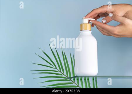 Donna corta e irriconoscibile che mostra una bottiglia cosmetica bianca con un tappo in legno, ideale per il branding di mockup, su uno sfondo blu con una l verde Foto Stock