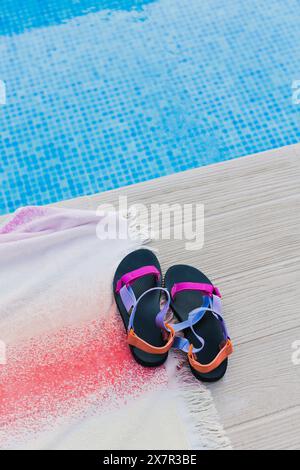 I sandali dai colori vivaci si trovano accanto a una scintillante piscina blu, arroccati su un ponte di legno testurizzato parzialmente ricoperto da un asciugamano bianco con frange cosparso Foto Stock