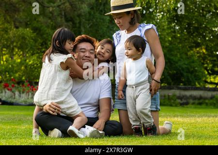 Una felice famiglia asiatica con tre bambini che abbracciano il padre in una giornata di sole in un parco verde durante una vacanza in Europa Foto Stock