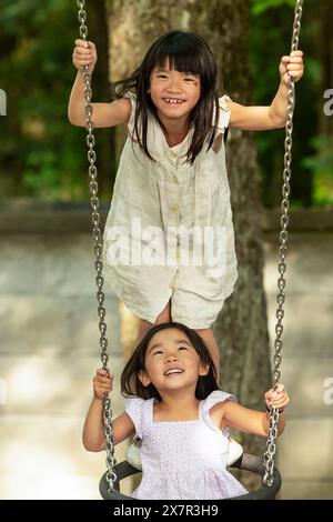 Due sorelle asiatiche si godono un momento di gioco su un altalena in un parco europeo, sorridendo e divertendosi insieme durante la loro vacanza in famiglia Foto Stock