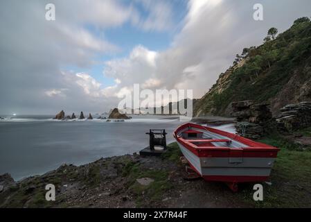 Un tranquillo paesaggio caratterizzato da formazioni rocciose costiere frastagliate e una barca rossa fissa sotto un dinamico cielo nuvoloso in Cantabria, Spagna Foto Stock