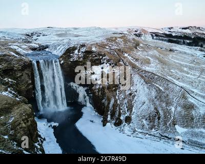 Una foto aerea mozzafiato di una potente cascata che sgorga tra scogliere innevate nel terreno accidentato dell'Islanda Foto Stock