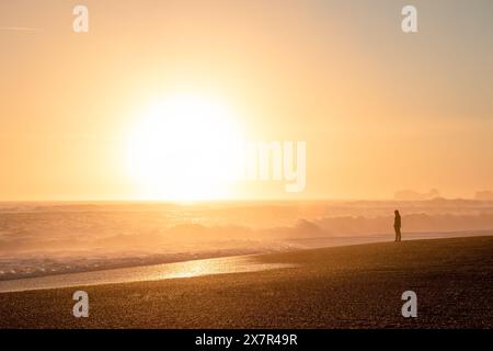 Un vibrante tramonto proietta un bagliore dorato su una spiaggia islandese, dove la silhouette di una persona solitaria viene catturata sullo sfondo di un oceano potente Foto Stock