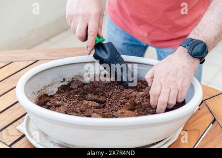 Vista ravvicinata delle mani di un giardiniere maschio utilizzando una cazzuola per mescolare terreno ricco in una grande piantatrice bianca su una superficie in legno Foto Stock