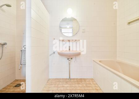 Bagno immacolato e ben illuminato con lavabo a parete, specchio circolare, pareti piastrellate e vasca e doccia combinate Foto Stock