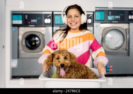Felice ragazza preadolescente con le cuffie in una lavanderia, in posa con un cane giocoso seduto in un cestino per la lavanderia, entrambi guardando la macchina fotografica. Foto Stock