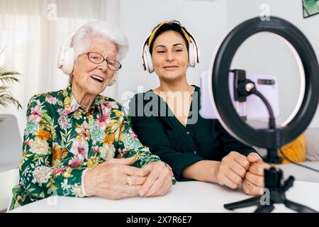 Un giovane influencer e una donna anziana sono seduti a una scrivania e registrano un podcast, entrambi indossano le cuffie e sorridono alla telecamera Foto Stock