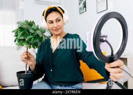 Una donna influencer registra un podcast a tema, spiegando la cura di un impianto e utilizzando una luce anulare per una qualità video perfetta Foto Stock
