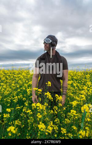Un uomo vestito da pilota d'epoca, che include occhiali e un casco in pelle, si erge in modo contemplativo in un campo vibrante di fiori gialli sotto a. Foto Stock