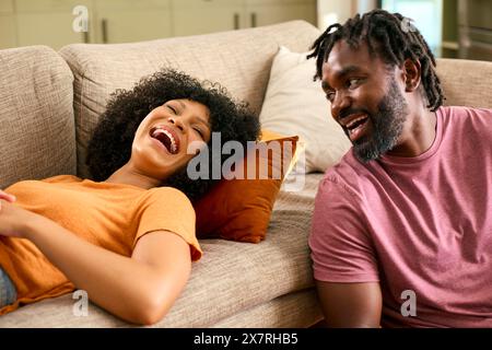 Coppia ridendo a casa con donna sdraiata sul divano e uomo seduto sul pavimento Foto Stock