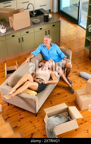 Foto della coppia matura in pausa dal disimballaggio sul divano con drink in New Home il giorno del trasloco Foto Stock