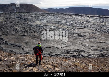 Turista donna con zaino in piedi sulla cima delle rocce di basalto fuso nel campo di lava del vulcano Fagradalsfjall, osservando il paesaggio vulcanico dell'Islanda. Foto Stock