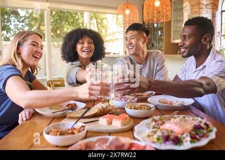 Gruppo di amici multirazziali seduti intorno al tavolo gustando un pasto a casa facendo saluti con l'acqua Foto Stock