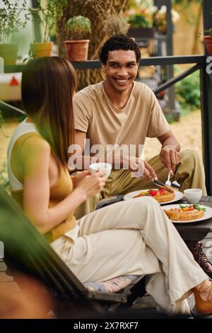 Una coppia interrazziale si diverte a fare un picnic su una panchina, condividendo un pasto insieme in un momento di romantico legame. Foto Stock