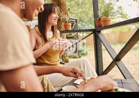 Una coppia interrazziale gode di un momento tranquillo su una panchina, condividendo un pasto insieme in un parco panoramico. Foto Stock