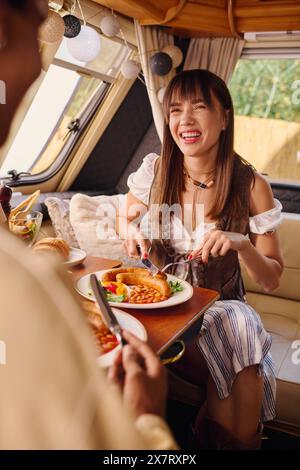 Una coppia interrazziale si gode un pasto sul retro di un camper durante la loro fuga romantica. Foto Stock