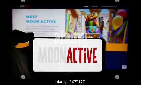 Persona che detiene un cellulare con il logo della società israeliana di giochi mobili Moon Active Ltd. Di fronte alla pagina Web aziendale. Mettere a fuoco il display del telefono. Foto Stock