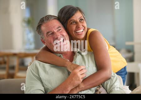 Coppia matura a casa con donna che abbraccia e uomo sorprendente seduto sul divano Foto Stock