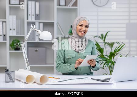 Architetto donna sicura di sé con hijab che utilizza un tablet in un ufficio moderno, circondato da documenti di design e un notebook. Foto Stock