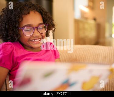 Ragazza che indossa gli occhiali a casa seduta sul divano che legge Una storia insieme Foto Stock
