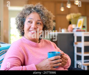 Donna matura seduta sul divano di casa, che si rilassa con una bevanda calda Foto Stock