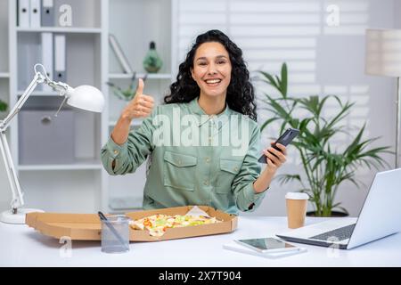 Una donna allegra che si gusta la pizza alla scrivania del suo ufficio, si regge un pollice e tiene in mano un telefono. Foto Stock