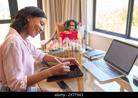 Madre che lavora da casa su un tablet digitale mentre la figlia ascolta musica e suona in sottofondo Foto Stock