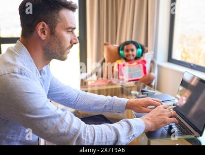 Padre che lavora da casa su un tablet digitale mentre la figlia ascolta musica e suona in sottofondo Foto Stock