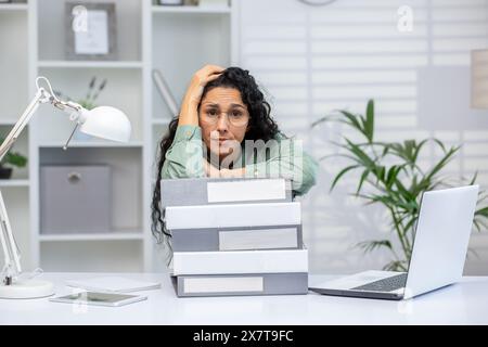 Donna stressata con gli occhiali sopraffatti al lavoro, seduta a una scrivania con una pila di leganti in un moderno ambiente d'ufficio. Foto Stock