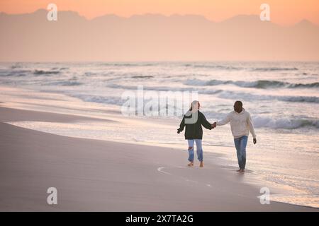 Una giovane coppia amorevole vestita in modo informale che cammina lungo il litorale della spiaggia tenendosi per mano all'alba Foto Stock