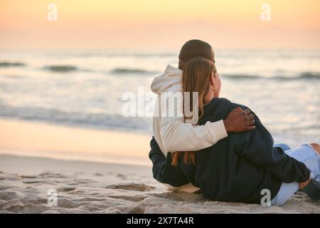 Vista posteriore di una giovane coppia amorevole, vestita in modo informale, seduta sulla spiaggia e abbracciata al sorgere del sole Foto Stock