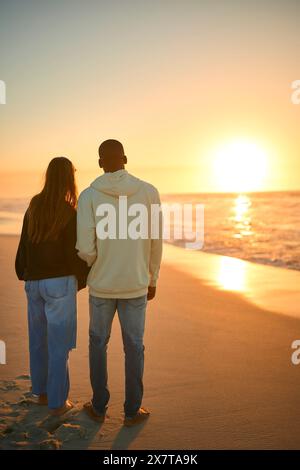 Vista posteriore di una giovane coppia amorevole in piedi sulla spiaggia che abbraccia all'alba Foto Stock
