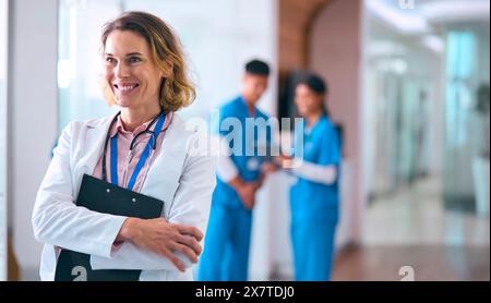 Donna Dottore in cappotto bianco con appunti in ospedale con colleghi in background Foto Stock