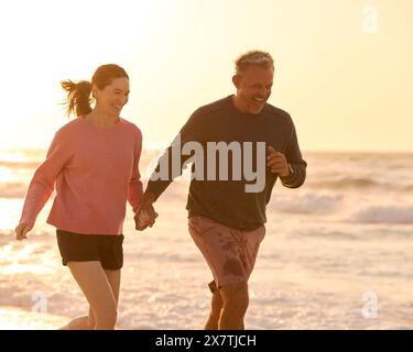 Una coppia anziana in pensione in vacanza che corre lungo il litorale della spiaggia tenendosi per mano al Sunrise Foto Stock
