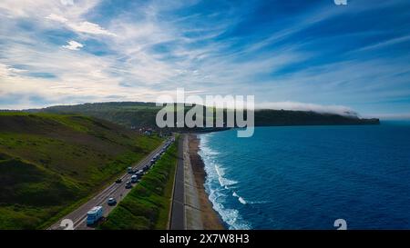 Una strada costiera che corre lungo una spiaggia con onde che si infrangono sulla riva. La strada è delimitata da verdi colline da un lato e dall'oceano dall'altro. Foto Stock