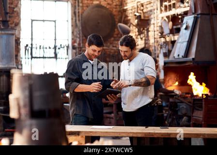 Uomini, fabbro e tablet in laboratorio sul lavoro di squadra per la ricerca e la collaborazione. Piccole imprese, artigianato e metallo con la navigazione su Internet per l'acciaio Foto Stock