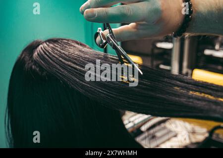 Il parrucchiere fa un acconciatura per donna . Parrucchiere uomo con una barba lunga in un gilet, esegue abilmente una acconciatura con un pettine e un taglio di capelli a una donna con lunghi capelli neri in un negozio di parrucchieri. Concetto di approccio al cliente di abilità. colpo medio Foto Stock