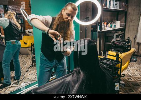 Il parrucchiere fa un acconciatura per donna . Parrucchiere uomo con una barba lunga in un gilet, esegue abilmente una acconciatura con un pettine e un taglio di capelli a una donna con lunghi capelli neri in un negozio di parrucchieri. Concetto di approccio al cliente di abilità. colpo medio Foto Stock