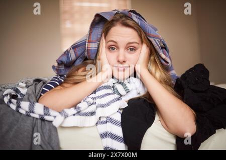 Servizio di pulizia, ritratto e donna con pila di lavanderia per le faccende domestiche, annoiata in camera da letto. Faccia, stanca e casalinga sul letto con vestiti sporchi e biancheria da letto Foto Stock